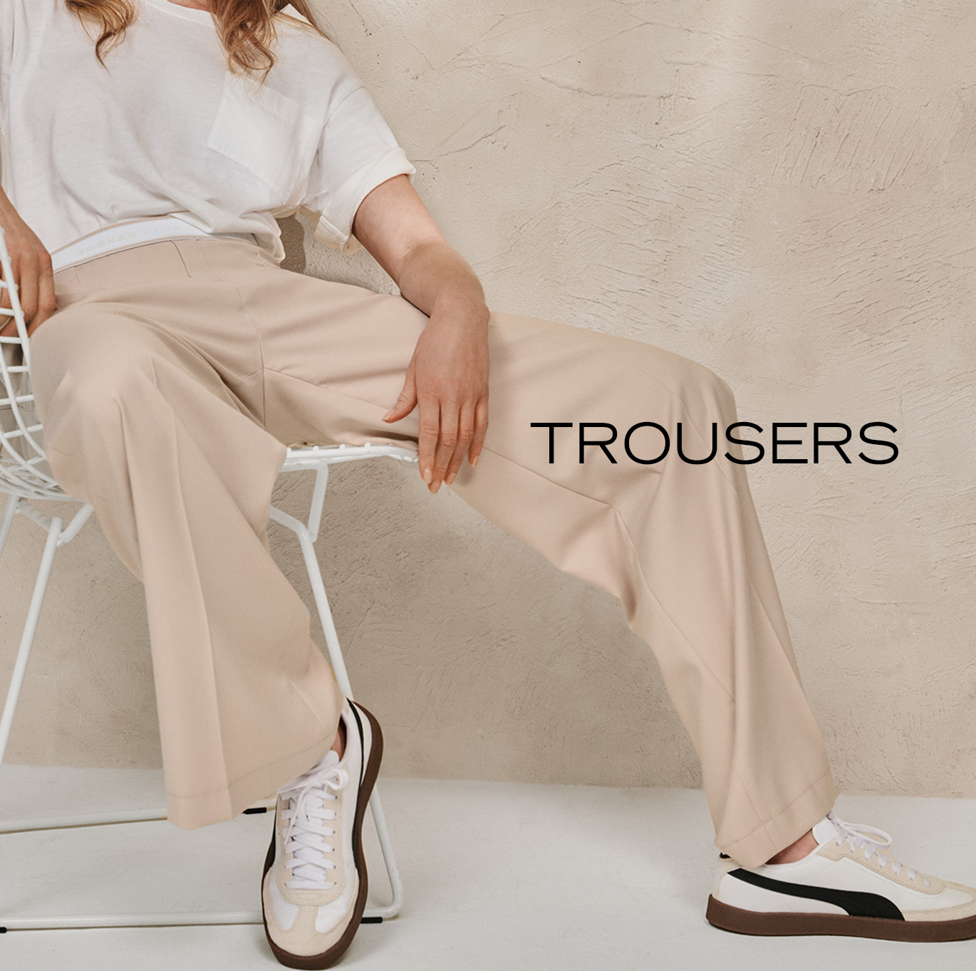 Model wearing wide-leg beige Hosen with a white T-shirt while seated on a chair – teaser for the ROSNER trousers category